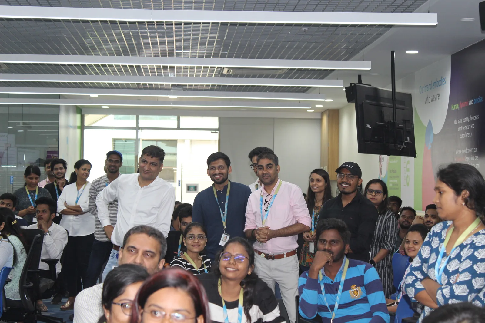 A packed Capgemini office floor, colleagues standing and seated, watching a session